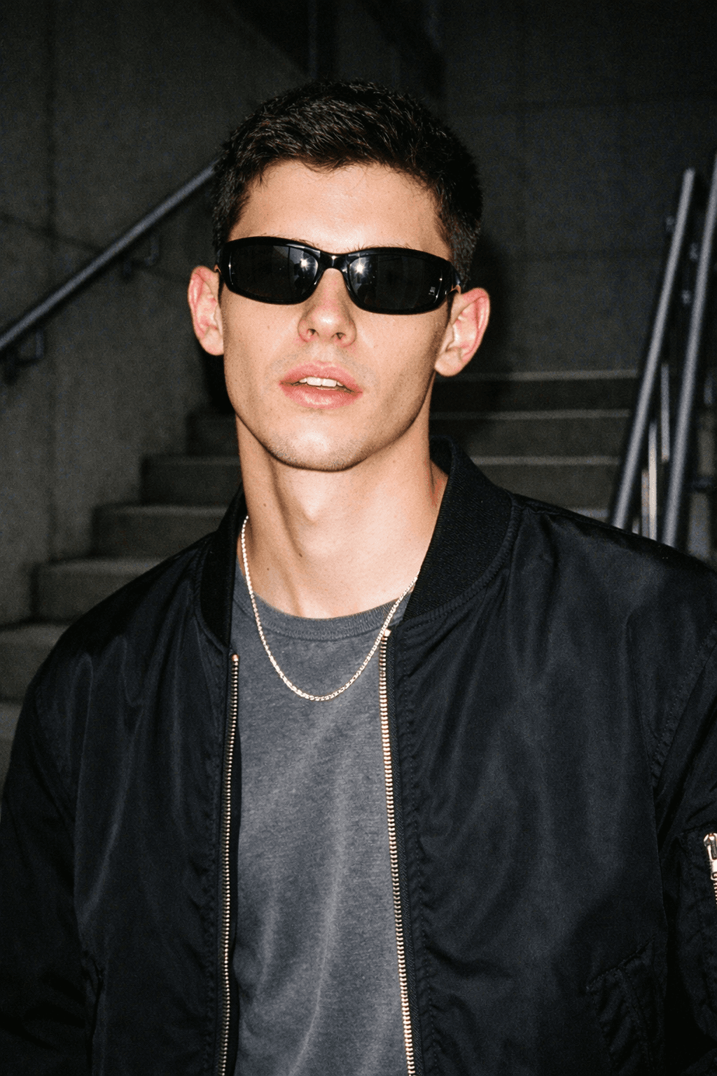 Close-up of a young male model wearing black Y2K sunglasses and a bomber jacket, photographed on a stairwell with harsh direct flash that brightens his face while the background falls into darker shadow.