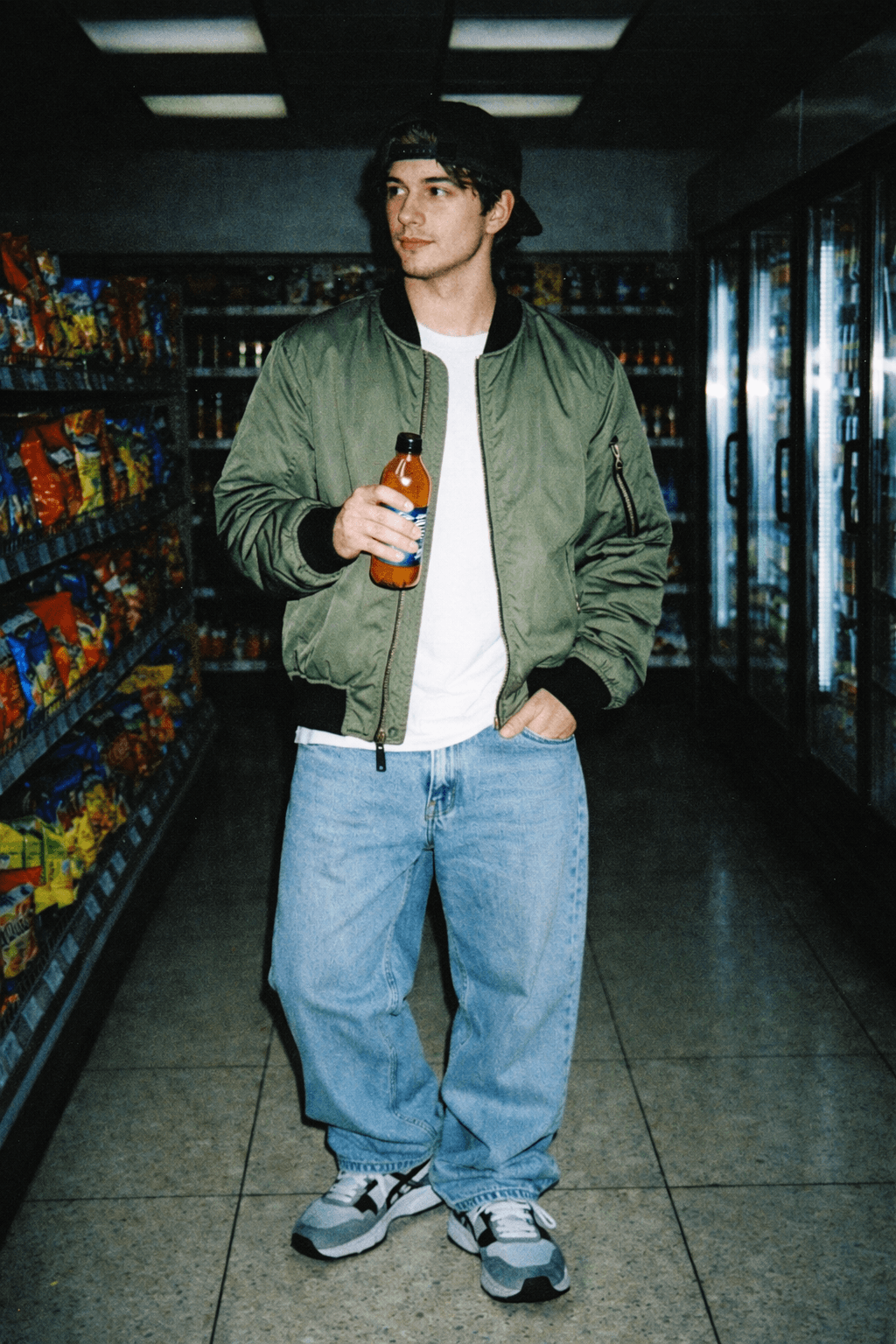 Young man standing in a convenience store aisle holding a drink bottle, lit by harsh disposable-camera flash that brightens him while the store behind falls into darker shadows.